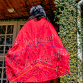 Person wearing a red garment with colorful patterns standing in front of a building with ivy.