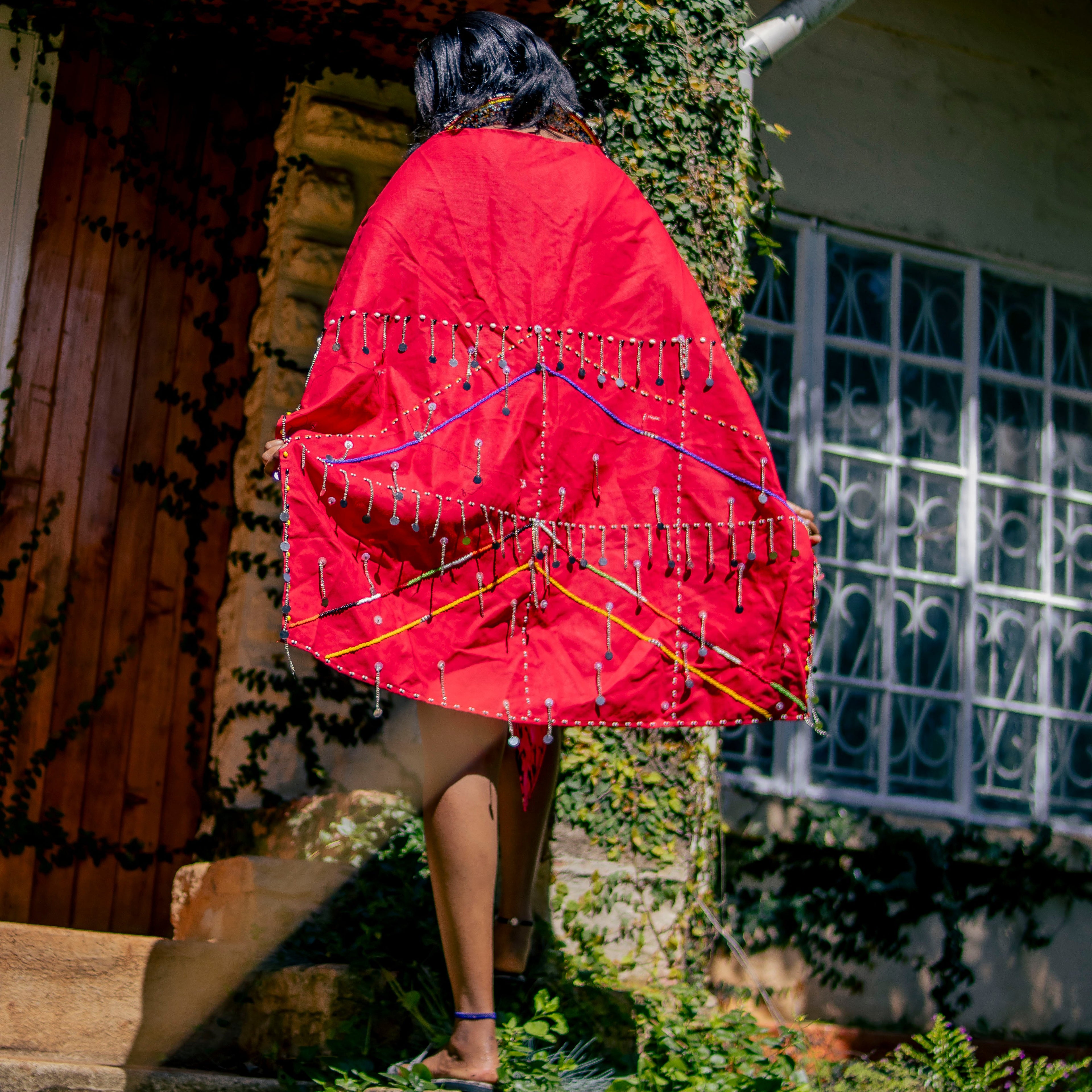 Person wearing a red garment with intricate patterns, standing in front of a building with a decorative metal grille.